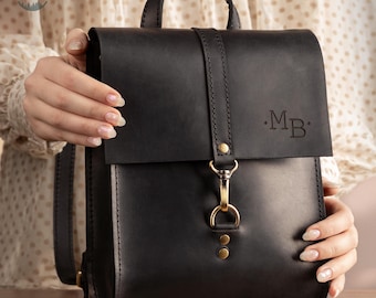 May include: A black leather backpack with a top handle and adjustable straps. The bag features a flap closure with a gold-colored clasp and two small rivets. The initials "MB" are embossed on the front. The text "Black S" is at the bottom.