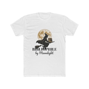 Puede incluir: Una camiseta blanca de manga corta con un gráfico de una bruja volando en una escoba mientras sostiene una pila de libros frente a una gran luna llena. Debajo del gráfico, el texto dice BIBLIOPHILE by Moonlight en letras negras.