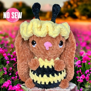 以下が含まれることがあります： ミツバチの衣装を着た、かぎ針編みで作ったぬいぐるみウサギ。ウサギは茶色で、黄色と黒のストライプ模様。ウサギはピンクの花畑の切り株に座っている。画像には「NO SEW CROCHET PATTERN」というテキストが表示されている。