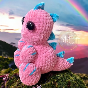 May include: A pink crocheted dinosaur toy with blue accents and large glittery eyes. The toy is sitting on a mossy rock with a rainbow in the background.