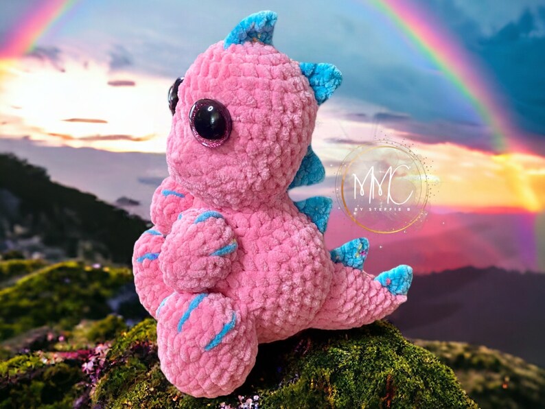 May include: A pink crocheted dinosaur toy with blue accents and large glittery eyes. The toy is sitting on a mossy rock with a rainbow in the background.