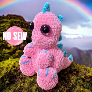 May include: Pink crocheted dinosaur toy with blue accents and large glittery eyes. The toy is sitting on a bed of green moss with a rainbow in the background. The text "NO SEW CROCHET PATTERN" is visible on a white circle.
