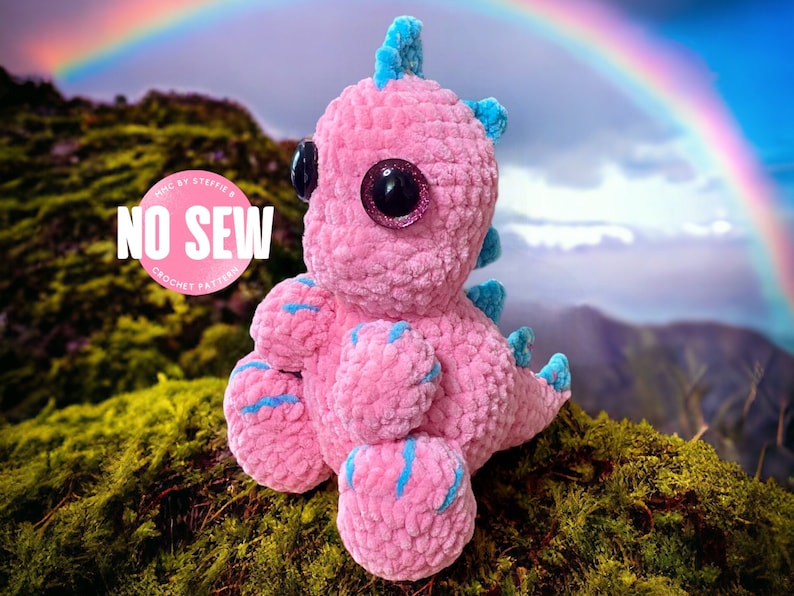 May include: Pink crocheted dinosaur toy with blue accents and large glittery eyes. The toy is sitting on a bed of green moss with a rainbow in the background. The text "NO SEW CROCHET PATTERN" is visible on a white circle.
