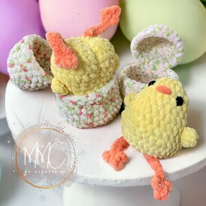 May include: Two crocheted yellow chicks with orange feet and beaks. One chick is sitting inside a crocheted egg with a floral pattern. The other chick is sitting on a white surface with a floral pattern.