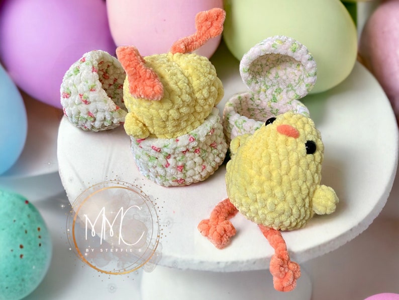 May include: Two crocheted yellow chicks with orange feet and beaks. One chick is sitting inside a crocheted egg with a floral pattern. The other chick is sitting on a white surface with a floral pattern.