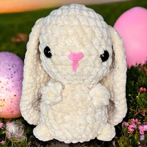 May include: A white crocheted bunny rabbit with pink nose and black eyes. The bunny is sitting on a bed of green grass with pink flowers and two Easter eggs.