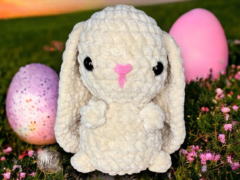 May include: A white crocheted bunny rabbit with pink nose and black eyes. The bunny is sitting on a bed of green grass with pink flowers and two Easter eggs.