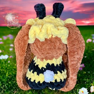 以下が含まれることがあります： 茶色、黄色、黒のストライプのクロシェ編みで作られた蜂のぬいぐるみ。蜂は長い茶色の耳と、黒い触角のある黄色の頭をしています。