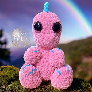May include: A pink crocheted dinosaur toy with blue accents and large black eyes. The toy is sitting on a bed of moss with a rainbow in the background.