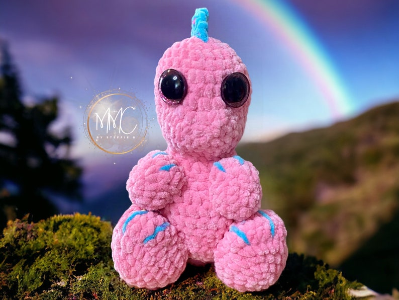 May include: A pink crocheted dinosaur toy with blue accents and large black eyes. The toy is sitting on a bed of moss with a rainbow in the background.