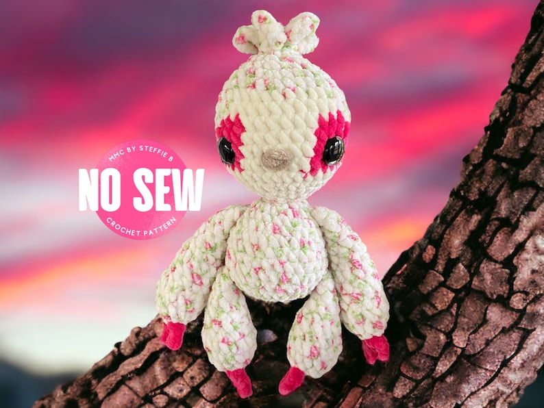 May include: A crocheted amigurumi toy in the shape of a creature with a pink and white floral pattern. The toy has large black eyes and pink accents. The toy is sitting on a tree branch against a pink and orange sunset background. The text "MHC by Steffie B NO SEW CROCHET PATTERN" is visible in the image.