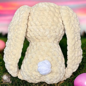May include: A crocheted plush bunny toy with long ears and a white fluffy tail. The bunny is cream colored and is sitting on a bed of green grass.
