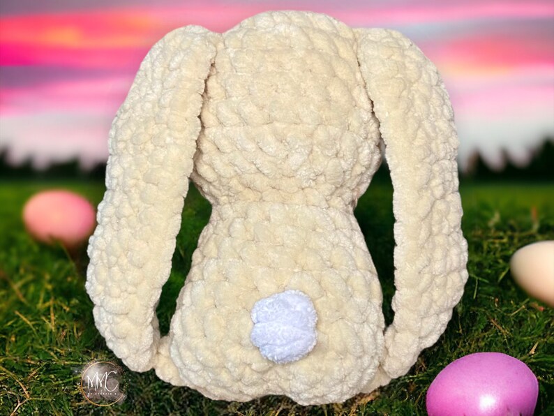 May include: A crocheted plush bunny toy with long ears and a white fluffy tail. The bunny is cream colored and is sitting on a bed of green grass.