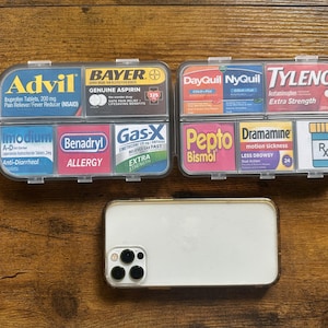 May include: Two clear plastic pill organizers filled with various over-the-counter medications, including Advil, Bayer, and Tylenol. A white smartphone with a gold-colored frame is below the organizers on a wooden surface.