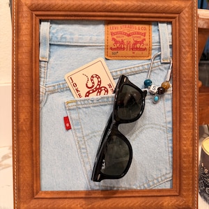 Może przedstawiać: Jasnoniebieskie tło z tkaniny dżinsowej w brązowej drewnianej ramie, z naszywką ze skóry Levi Strauss & Co., kartą do gry Joker z ilustracją węża, czarnymi okularami przeciwsłonecznymi i metalowym karabińczykiem z kolorowymi koralikami.