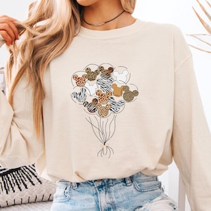 A woman wearing a white long-sleeved shirt with a graphic of a bunch of balloons and a zebra design, paired with denim shorts, sits on a bed with a pillow featuring a black and white eye design.