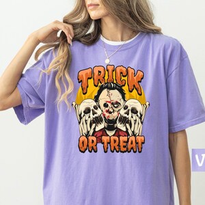 May include: Violet-colored t-shirt with a Halloween-themed graphic. The design features a scary character with two skulls and the words "Trick or Treat" in orange. The shirt is a comfortable, oversized fit.