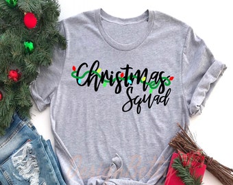 May include: Light gray t-shirt with the words "Christmas Squad" in black script, accented with a string of colorful Christmas lights. The shirt is paired with blue jeans and festive holiday decorations.
