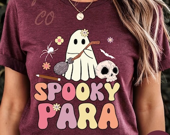 May include: Heather maroon t-shirt with a colorful graphic design. The design features a ghost with a flower, a broom, a skull, and the words "SPOOKY PARA" in a retro font. Additional elements include a spider, flowers, and a bat.