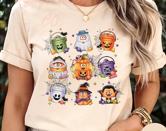 May include: Beige t-shirt featuring a grid of Halloween-themed characters. The characters include a ghost, pumpkin, mummy, and vampire, each with a unique design. The shirt has short sleeves and a round neckline. The word "NATURAL" is printed on a round sticker.