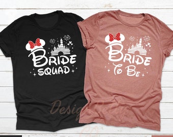 May include: Two t-shirts with a white graphic of a Disney castle, fireworks, and a Mickey Mouse ear headband. The text on the black shirt reads "Bride Squad" and the text on the heather mauve shirt reads "Bride to Be".