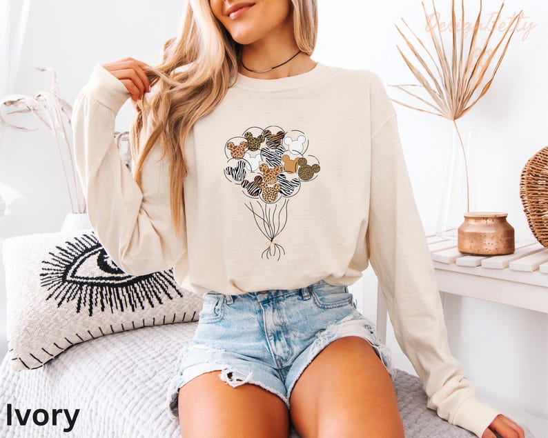 A woman wearing a white long-sleeved shirt with a graphic of a bunch of balloons and a zebra design, paired with denim shorts, sits on a bed with a pillow featuring a black and white eye design.