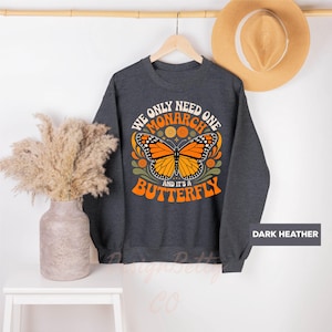 A gray sweatshirt with a graphic design of a butterfly and the text "We only need one monarch butterfly and it's a butterfly" is hanging on a wooden hanger. Next to the sweatshirt is a beige hat and a white vase with dried flowers.