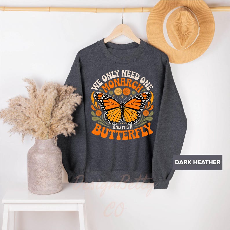 A gray sweatshirt with a graphic design of a butterfly and the text "We only need one monarch butterfly and it's a butterfly" is hanging on a wooden hanger. Next to the sweatshirt is a beige hat and a white vase with dried flowers.