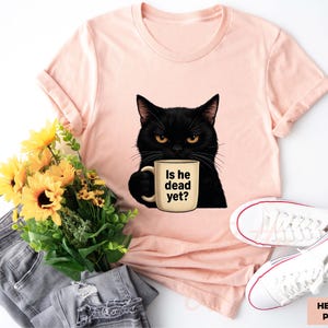 K&ouml;nnte beinhalten: Ein pinkfarbenes T-Shirt mit einer schwarzen Katze, die eine Kaffeetasse mit der Aufschrift "Is he dead yet?" h&auml;lt. Die Katze hat gelbe Augen und einen ernsten Gesichtsausdruck. Das Shirt liegt auf einem wei&szlig;en Hintergrund mit einem Paar wei&szlig;en Sneakers und einer grauen Jeans.