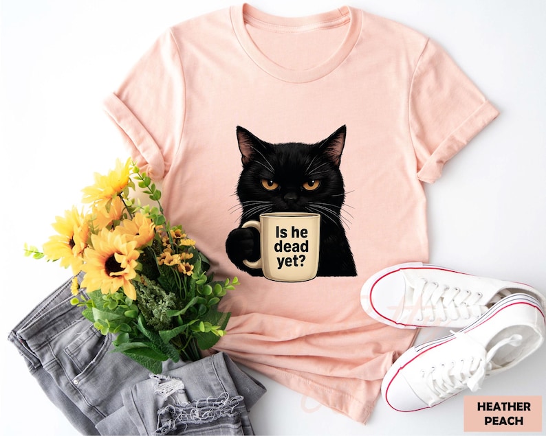 K&ouml;nnte beinhalten: Ein pinkfarbenes T-Shirt mit einer schwarzen Katze, die eine Kaffeetasse mit der Aufschrift "Is he dead yet?" h&auml;lt. Die Katze hat gelbe Augen und einen ernsten Gesichtsausdruck. Das Shirt liegt auf einem wei&szlig;en Hintergrund mit einem Paar wei&szlig;en Sneakers und einer grauen Jeans.
