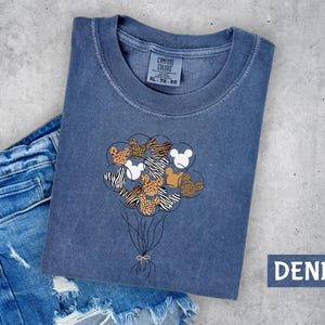 A blue denim t-shirt with a graphic design of a bunch of balloons featuring Mickey Mouse and other cartoon characters is displayed on a gray surface.