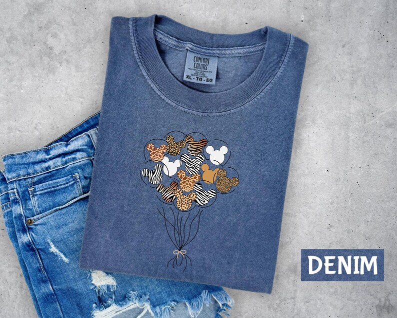 A blue denim t-shirt with a graphic design of a bunch of balloons featuring Mickey Mouse and other cartoon characters is displayed on a gray surface.