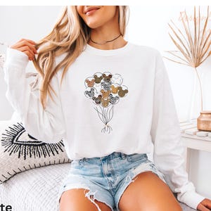 A woman wearing a white long-sleeved shirt with a graphic of a bunch of balloons and a choker necklace is sitting on a bed.