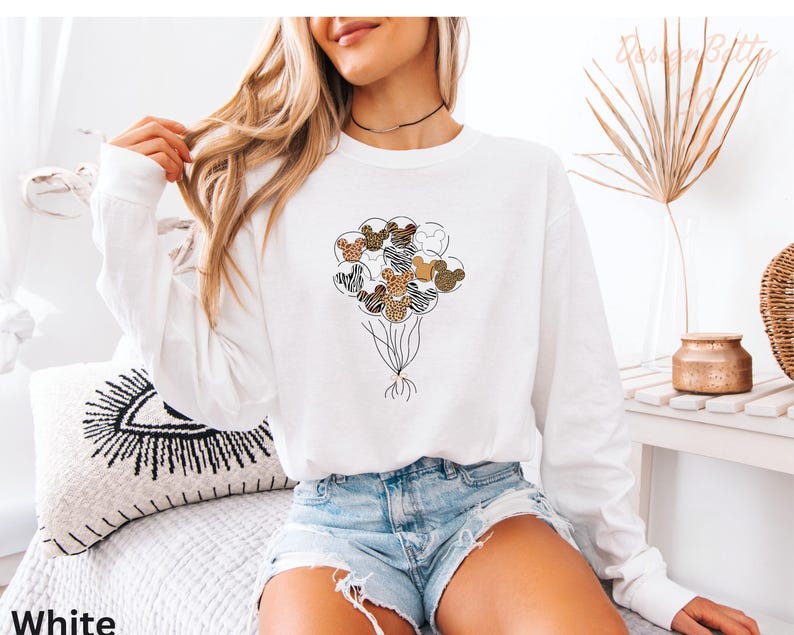 A woman wearing a white long-sleeved shirt with a graphic of a bunch of balloons and a choker necklace is sitting on a bed.