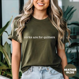 May include: Heather olive green t-shirt with rolled sleeves. The white text "corks are for quitters" is printed across the chest. The shirt is paired with blue jeans. The bottom right corner of the image has the text "HEATHER OLIVE".