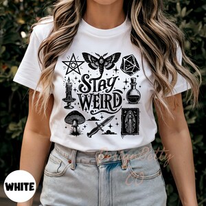 May include: White t-shirt with a black graphic design featuring a pentagram, moth, dice, candle, mushroom, potion bottle, dagger, and a framed image of a saint. The words "Stay Weird" are in the center.