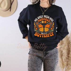 A person wearing a black sweatshirt with a graphic design of a butterfly and text, standing in front of a wall with a hat and a plant.