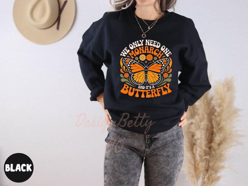 A person wearing a black sweatshirt with a graphic design of a butterfly and text, standing in front of a wall with a hat and a plant.