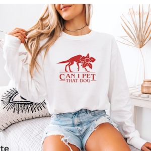 A woman wearing a white long-sleeved shirt with a red graphic and text printed on it, sitting on a bed with a decorative pillow.