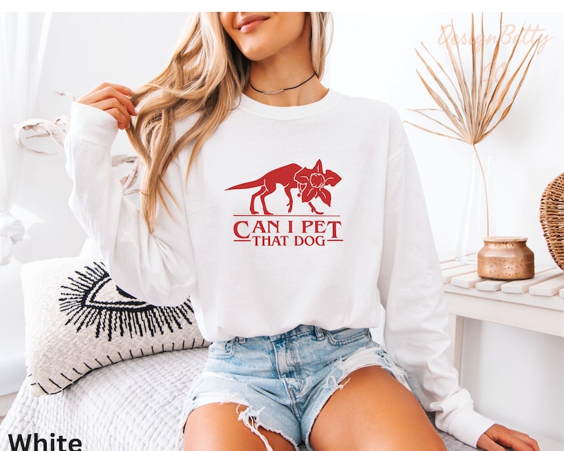 A woman wearing a white long-sleeved shirt with a red graphic and text printed on it, sitting on a bed with a decorative pillow.
