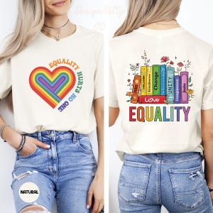 May include: Cream-colored t-shirt with a rainbow heart graphic and the text "EQUALITY HURTS NO ONE." The back of the shirt features a design of colorful books with words like "Love," "Kindness," and "Equality."