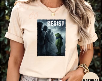 May include: Beige t-shirt with a graphic depicting a frog in a blue scarf facing riot police, with the word "RESIST" above. The shirt has short sleeves. The image has a political theme.