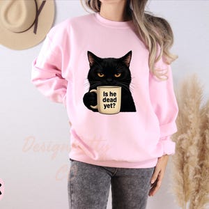 K&ouml;nnte beinhalten: Rosafarbener Pullover mit einer schwarzen Katze, die eine Kaffeetasse mit dem Text "Is he dead yet?" h&auml;lt.