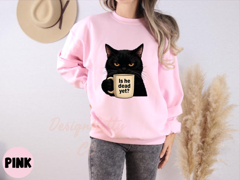 K&ouml;nnte beinhalten: Rosafarbener Pullover mit einer schwarzen Katze, die eine Kaffeetasse mit dem Text "Is he dead yet?" h&auml;lt.