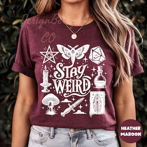 May include: Maroon t-shirt with a white graphic design featuring a moth, pentagram, dice, potion bottle, mushroom, dagger, and the words "Stay Weird". The shirt is made of a soft material.