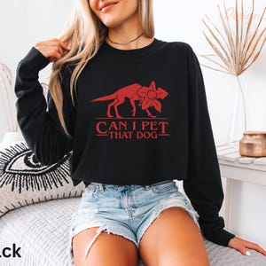 A woman wearing a black long-sleeved shirt with a red graphic and text printed on it, sitting on a bed with a pillow.