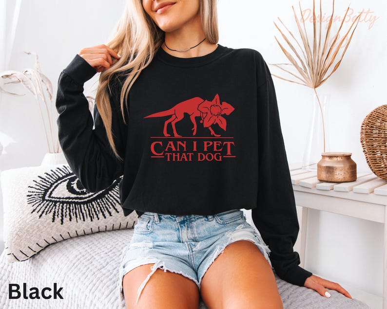 A woman wearing a black long-sleeved shirt with a red graphic and text printed on it, sitting on a bed with a pillow.