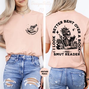 May include: Heather peach-colored t-shirt with a skeleton reading a book design. The front features a skeleton hand opening a book with the text "SPREAD THOSE PAGES." The back says "I LOOK BETTER BENT OVER A BOOK" and "PROUD SMUT READER."