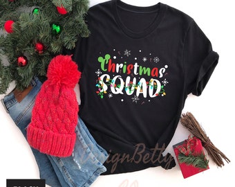 May include: Black crew neck t-shirt with the text "Christmas Squad" in white, green, and red. The design includes Christmas lights, snowflakes, and candy canes. A red knit hat and blue jeans are next to the shirt.