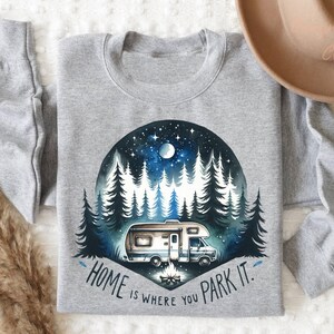 May include: A gray sweatshirt with a watercolor illustration of a camper van parked in a forest at night. The text "Home is where you park it." is printed below the illustration.